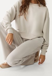 Jasno beżowy sweatshirt i pasujące dresy wykonane z miękkiego materiału. Sweatshirt ma okrągły dekolt i mankiety na rękawach, podczas gdy dresy mają luźny krój.