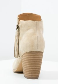 Beige suède enkellaars met een ronde neus, franjes, een rits aan de zijkant en een gestapelde houten hak met horizontale groeven.