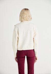 Suéter de punto crema con dobladillo acanalado y botones en los hombros, combinado con pantalones burdeos. Enfóquese en la textura y el contraste de colores.