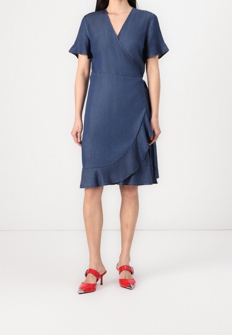 Denim wrap dress yang menampilkan lengan pendek, leher V, hem berumbai, dan belahan di samping. Dipasangkan dengan sepatu hak tinggi merah dengan aksen metalik.