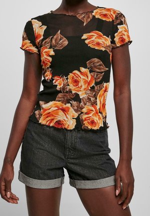 Vrouw die een zwarte doorschijnende bloementop met oranje rozen draagt en donkere denim shorts met opgerolde zoom, staand tegen een effen achtergrond.