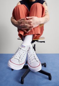 Scarpe da ginnastica basse bianche con dettagli rossi e suole in gomma. Indossate con calzini bianchi e pantaloni arancioni, seduto su una sedia.