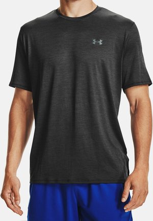 Mężczyzna ubrany w ciemnoszarą sportową koszulkę z krótkim rękawem z małym logo na piersi oraz niebieskie spodenki.