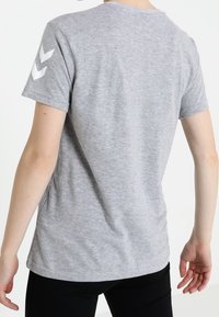 Camiseta de manga corta gris de algodón suave con cuello redondeado, que presenta acentos en forma de Chevron blancos en el hombro izquierdo.