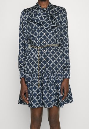 Femme portant une robe bleu marine à manches longues avec un motif chaîne blanc, ourlet à volants, boutons sur le devant, col à nœud et ceinture fine imprimé léopard.