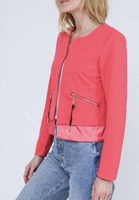 Veste cropped corail avec un col rond, des manches longues et des poches avant zippées. Présente une texture lisse et un ourlet en satin contrasté.