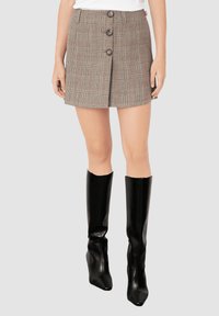 Mujer vestida con una falda corta de cuadros marrón y botones en la parte delantera, y botas de cuero negras hasta la rodilla, contra un fondo claro y liso.