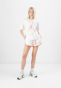 T-shirt bianco in cotone con logo floreale di Jordan, abbinato a pantaloni corti bianchi con rifiniture arancioni e logo. Le scarpe sono bianche con dettagli verdi.