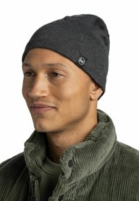 Gray ribbed knit beanie with a logo patch, worn by a person in a green corduroy jacket. Beanie has a slouchy silhouette.