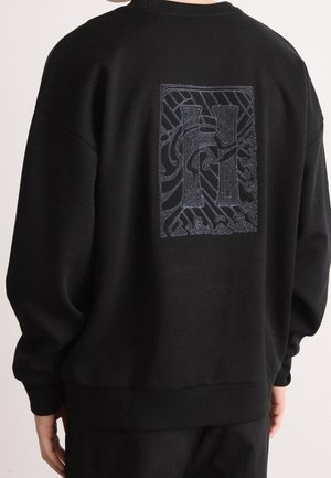 Person, der bærer en sort sweatshirt med et stort, udsmykket mørkegråt bogstav "H" design på den øverste del af ryggen.