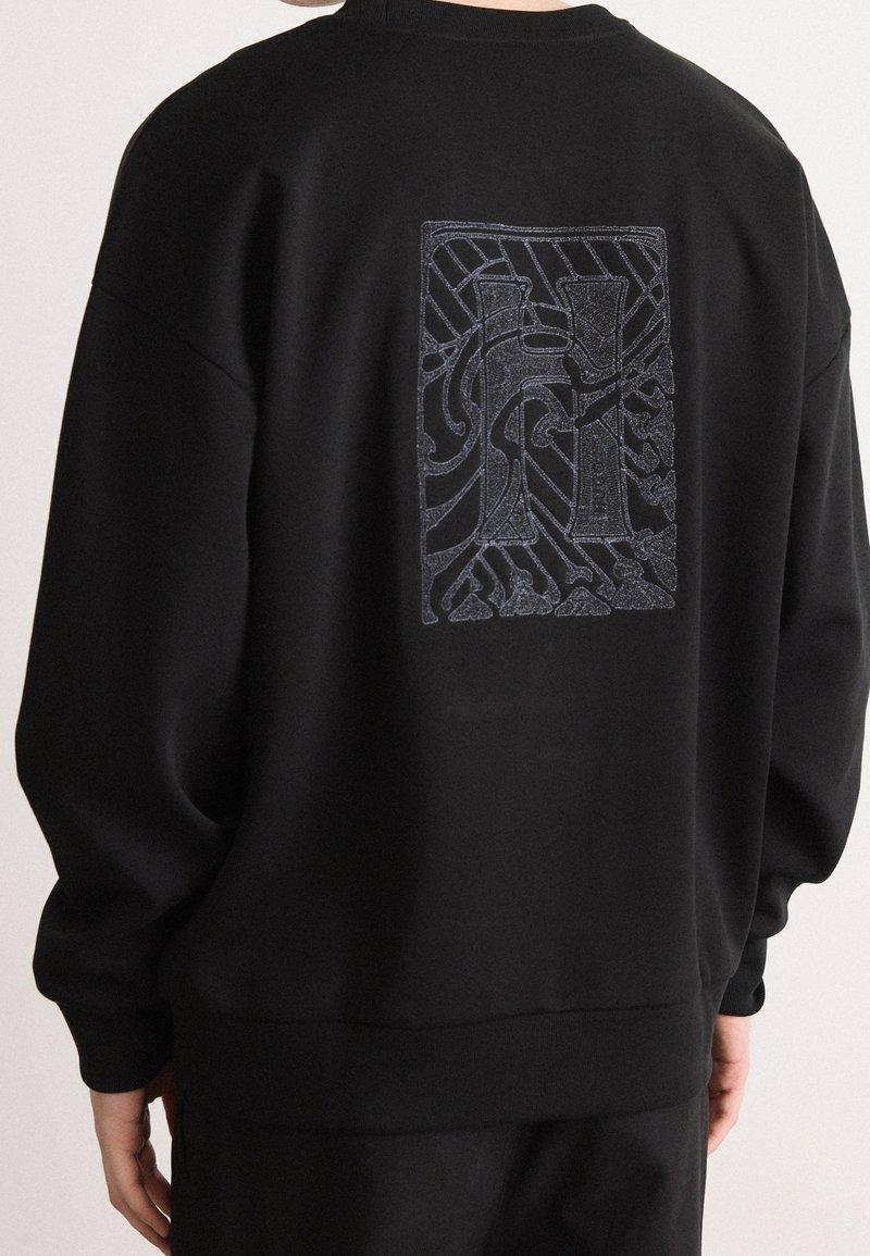 Personne portant un sweat-shirt noir avec une grande lettre ornée gris foncé "H" sur le haut du dos.
