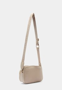 Borsa crossbody rettangolare beige realizzata in materiale morbido, dotata di una robusta tracolla con un logo in rilievo. Chiusura con zip in cima.