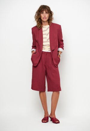 Vrouw met krullend haar die een bordeauxkleurige blazer en knielange shorts draagt, een beige gestreepte top en bijpassende bordeauxkleurige sandalen met hak.