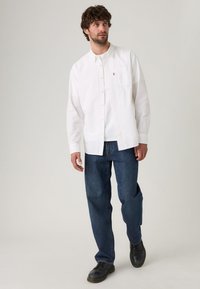 Camisa blanca de botones con cuello, un bolsillo en el pecho y el logo de Levi's; combinada con jeans oscuros y zapatos negros.