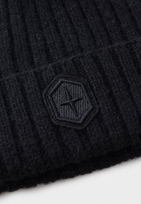 Black knitted beanie with vertical ribbing, featuring a hexagonal black leather patch embossed with a star design. Soft texture.