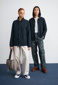 Deux mannequins se tiennent sur un tapis bleu. L'un porte une veste sombre, un pantalon clair et des baskets noires, tenant un sac gris. L'autre arbore une veste sombre et un pantalon à motifs avec des chaussures marron.