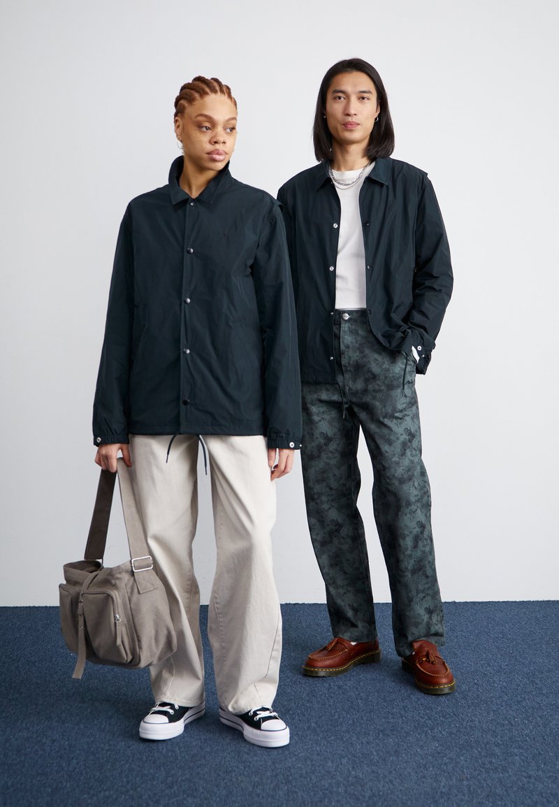 Due modelli si trovano su un tappeto blu. Uno indossa una giacca scura, pantaloni chiari e sneaker nere, mentre tiene in mano una borsa grigia. L'altro indossa una giacca scura e pantaloni a fantasia con scarpe marroni.