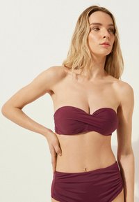 Bikini top sans bretelles de couleur bordeaux foncé avec un design torsadé ; bas taille haute assortis avec une texture froncée sur les côtés. Finition en tissu lisse.