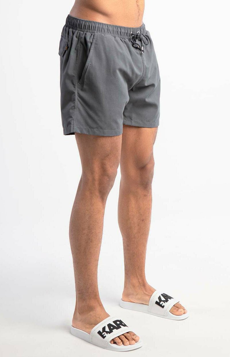 Man in grijze elastische taille shorts en witte slidesandalen met zwarte letters, staand op een witte achtergrond.