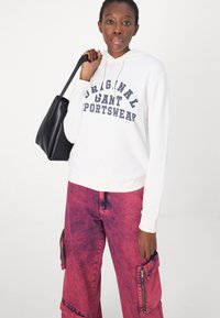Sweat à capuche blanc avec le graphique "Original GANT Sportswear" en marine, associé à un pantalon cargo rose, déchiré, avec des zips sur les côtés. Sac fourre-tout noir.