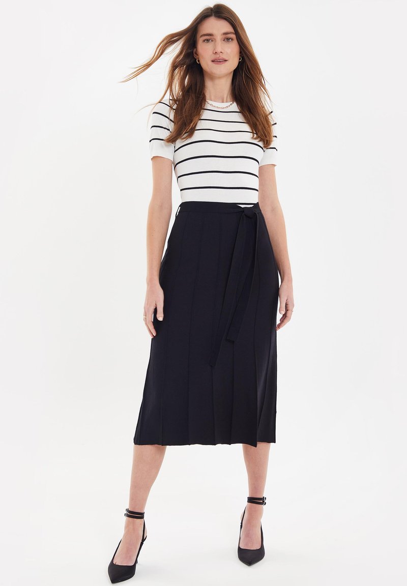 Top a maniche corte a righe in bianco e blu navy, abbinato a una gonna midi plissettata blu navy con vita legata e scarpe con tacco nere.