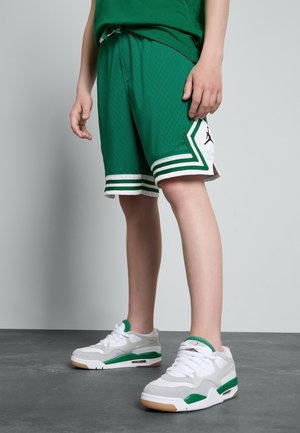 Osoba ubrana w zielone szorty do koszykówki z białą i zieloną lamówką oraz białe buty sportowe z zielonymi akcentami stojąca na szarej podłodze.