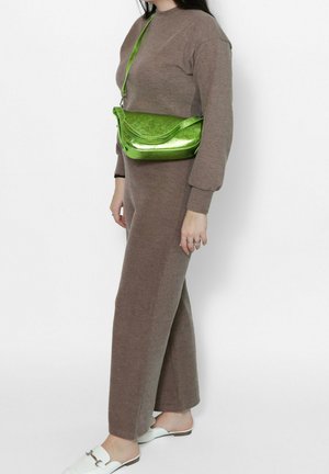 Donna che indossa un vestito marrone a maglia e scarpe slip-on bianche, con una lucida borsa verde a tracolla portata sul petto.