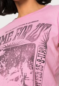 Camiseta de algodón rosa que presenta una gran impresión gráfica negra con texto e imágenes estilizadas, y un cuello redondo. Textura suave y corte relajado.