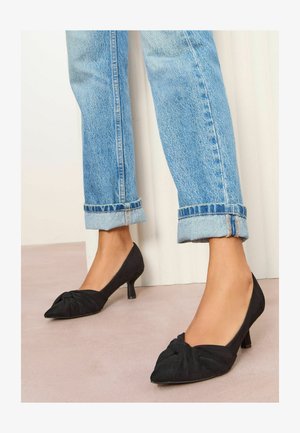 Schwarze Wildlederschuhe mit spitzer Zehe und Knoten-Detail an der Vorderseite, kombiniert mit hochgekrempelten hellblauen Jeans. 7,5 cm Absatz.