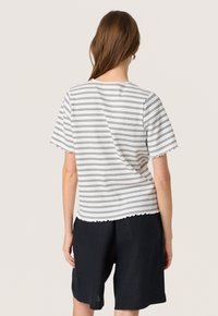 Mujer con cabello largo castaño, vistiendo una camiseta blanca de manga corta a rayas y pantalones cortos negros hasta la rodilla, vista desde atrás.