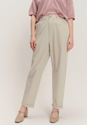 Donna che indossa pantaloni beige a vita alta con gamba dritta e orli risvoltati, abbinati a scarpe Mary Jane con tacco blocco color nude e una maglietta rosa larga.