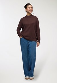 Maglione marrone con collo alto, abbinato a jeans blu a gamba larga e sneakers bianchi con suola marrone.