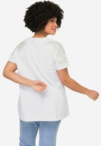 Weißes T-Shirt mit kurzen Ärmeln und verzierten silbernen Blumenakzenten an den Schultern, mit lockerer Passform und weichem Baumwollstoff.