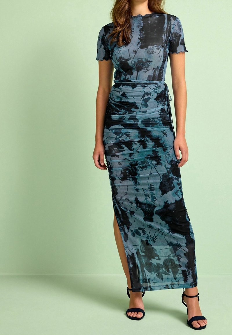 Femme portant une robe maxi cintrée à manches courtes, à motifs floraux bleus et noirs, avec une fente latérale et des talons ouverts noirs.