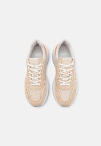 Beige idrottsskor i mocka och mesh-material, vita snören, texturerade detaljer och orange accent på hälen.