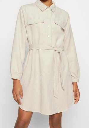 Helles beiges Corduroy-Hemdkleid mit langen Ärmeln, Knopfleiste, zwei Brusttaschen und einem Stoffgürtel, der auf der Taille gebunden ist.