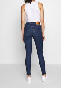 Jeans ajustados de mezclilla de color azul oscuro, con bolsillos traseros estándar y una etiqueta de marca de cuero marrón en la cintura. Se llevan zapatillas blancas.