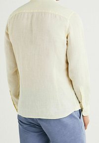 Camisa de manga larga amarilla clara hecha de lino texturizado, con cuello clásico y puños abotonados, vista desde atrás.