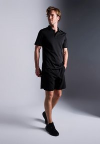 Μαύρο πόλο μπλουζάκι και σορτς από ελαφρύ ύφασμα, με απλό σχέδιο. Φορεμένα με μαύρα αθλητικά παπούτσια, σε γκρι φόντο.