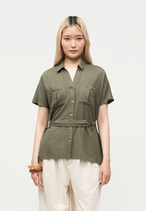 Young woman with long blonde hair wearing an olive green belted short-sleeve shirt and loose white pants against a neutral background.