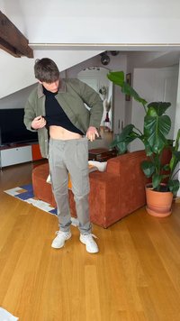 Jeune homme en vêtements décontractés ajustant la taille de son pantalon dans un salon avec un canapé orange, une grande plante en pot et un sol en bois.