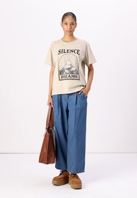 Beige grafisk t-skjorte med "SILENCE DREAMS" trykk, kombinert med løse blå denimbukser og brune platåsko, holder en brun totebag.