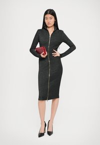 Rochie neagră, adaptată, dintr-un material texturat cu fermoar zarioasă în față, mâneci lungi și design până la genunchi. Modelul ține o poșetă roșie.
