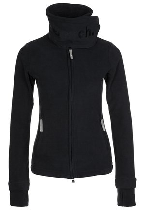 Kvinde gående iført en lilla t-shirt, mørk jakke, blå jeans, sorte sneakers og bærende en sort/hvid mønstret skuldertaske.