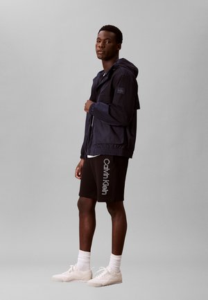 Joven con chaqueta oscura con capucha, pantalones cortos negros de Calvin Klein, calcetines blancos y zapatillas blancas, de pie frente a un fondo gris liso.