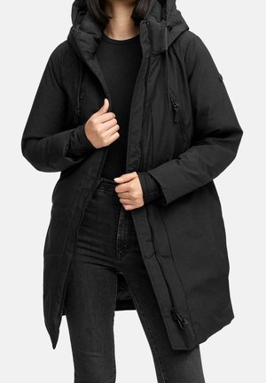 Femme portant un manteau d'hiver noir à capuche, partiellement zippé, par-dessus un haut noir et un jean noir, debout devant un fond uni.