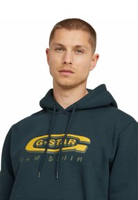 Ciemnozielona bluza z kapturem wykonana z mieszanki bawełny, z dużym żółtym logo G-Star i napisem "RAW DENIM". Posiada sznurki do regulacji i kieszeń kangurką.