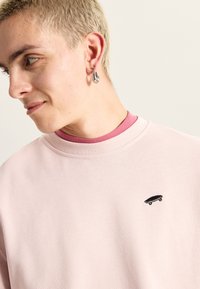 Lyserød sweatshirt med rund hals, der har et lille sort skateboardlogo på brystet. Glat tekstur og afslappet pasform.