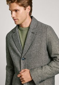 Blazer gris de espiga con un solo botón, solapas de muesca y un ajuste entallado. Lleva puesto un suéter verde. Detalle de tejido texturizado visible.