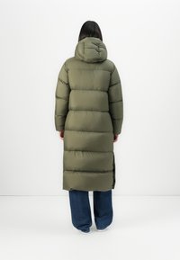 En person står med ryggen til iført en lang, olivengrøn, polstret frakke med hætte, blå jeans og hvide sko foran en ensfarvet baggrund.